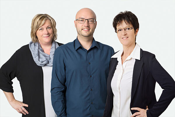 Drei freundliche Mitarbeiter eines Unternehmens stehen gemeinsam vor hellem Hintergrund.